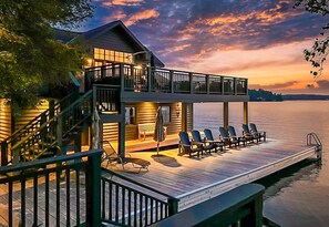 A waterfront property with a wooden deck, outdoor seating, and a covered area with a bench and umbrella. - Hard packed Sandy Beach, spacious 7 brdm 6 bath! Boathouse w Sleeping & Sundeck! (Port Carling)