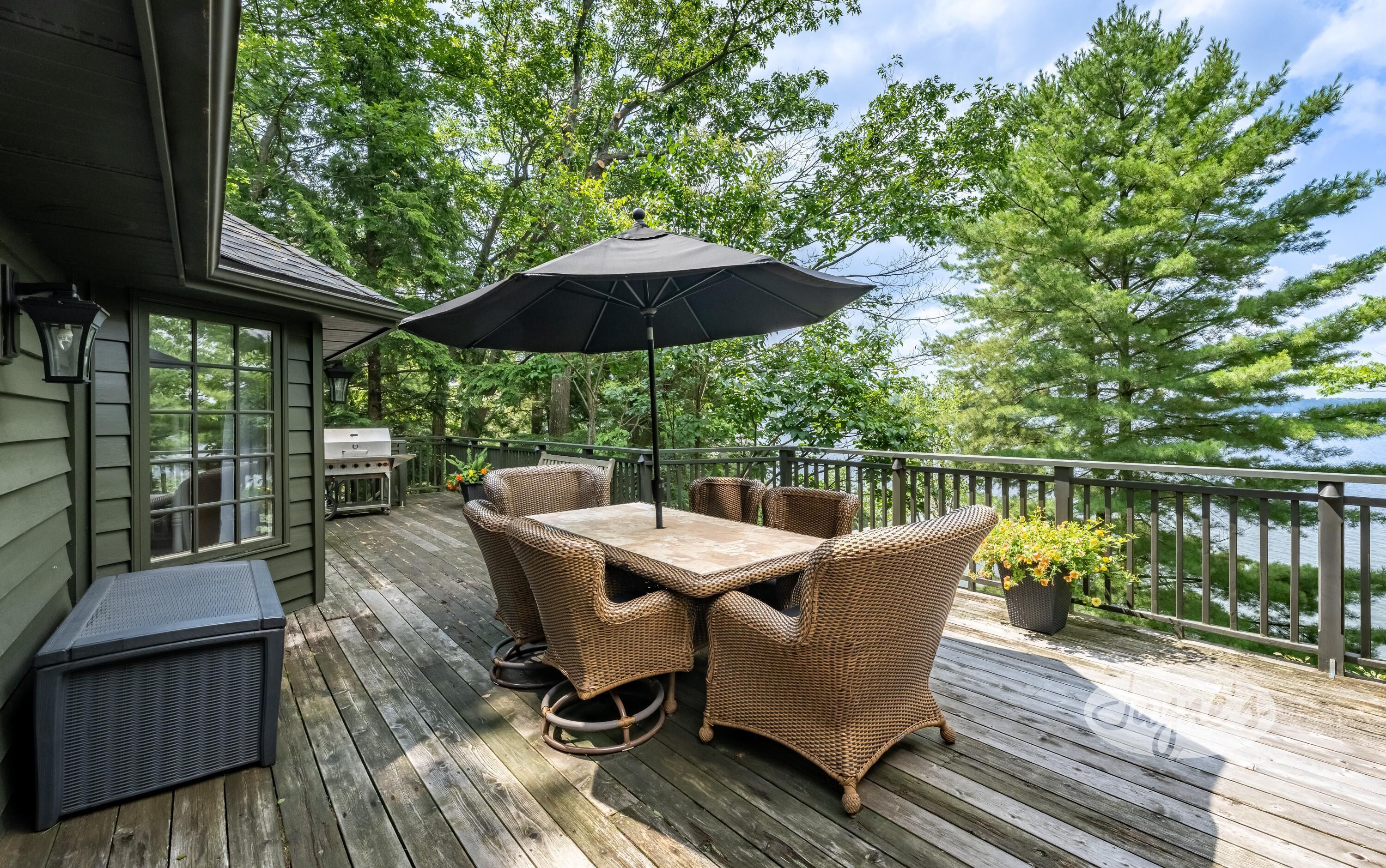 Una terraza de madera con mobiliario exterior, una parrilla y vistas a árboles y agua.