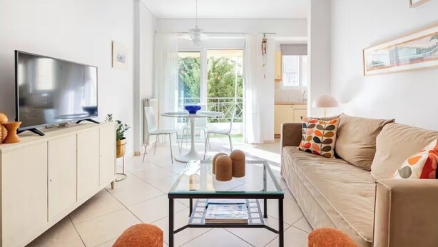 A modern living room with a TV, glass coffee table, sofa, and a balcony with a dining area. - Sunny Apartment Steps from Sepolia Metro (Athina)