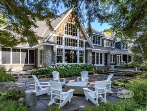 A large, two-story house with a stone facade, multiple windows, and a stone patio with Adirondack chairs. - 6 bed, 8 bath Beautiful Lake Joe Compound! Sunsets, Boathouse, Sauna & Gym! (Port Carling)