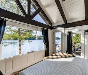 A room with a large window overlooking a lake, a bed with a patterned pillow, and a view of the outdoors. - Priceless views at this 6 bed, 5 bath Cottage + 1 bed Bunkie (Rosseau)
