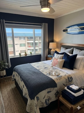 A hotel room with a bed, a window with a view of buildings, a ceiling fan, and a lamp on a bedside table.