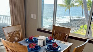 Una mesa de madera con cuatro sillas, adornada con platos azules y flores rojas, cerca de una ventana con vista al océano y palmeras.