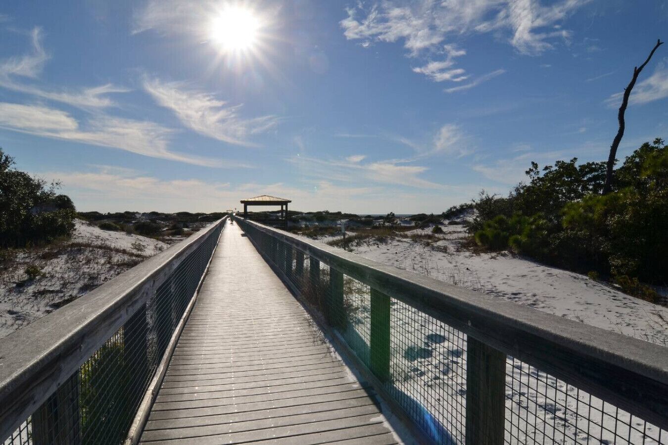 A wooden boardwalk leading to a covered structure, surrounded by sand dunes and sparse vegetation.