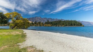 Vista al lago con una playa de guijarros, un árbol grande y una cadena montañosa al fondo.