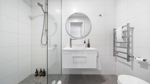 Een moderne badkamer met een glazen douchecabine, een ronde spiegel, een witte wastafel met een zeepdispenser en een handdoekhouder.