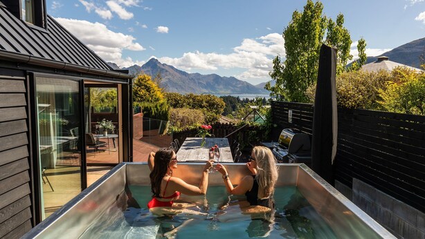 Un jacuzzi con dos personas, un patio con mesa y sillas, y vista a la montaña.