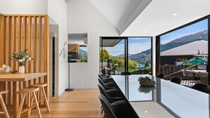 Un comedor moderno con una mesa larga y sillas, un separador de madera, y vistas a montañas y un lago a través de amplios ventanales.