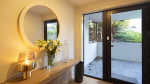 Zona de recepción de hotel con un espejo redondo, un jarrón de flores, una lámpara y un panel de vidrio corredera que da a un balcón con vegetación.