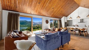 A spacious living room with a large window offering a scenic mountain view, a cozy seating area with a blue sofa and leather chairs, and a wooden ceiling. - Sicilian Luxury Retreat 2 | Alfresco Dining | Close to Wineries (Queenstown)