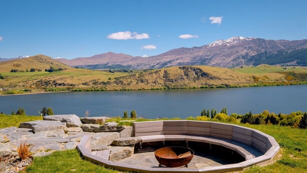 A circular stone fire pit with a metal bowl, surrounded by a stone pathway and grass, overlooking a lake with mountains in the background. - Sicilian Luxury Retreat 2 | Alfresco Dining | Close to Wineries (Queenstown)