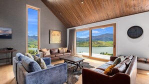 A spacious living room with large windows offering a scenic mountain view, a wooden ceiling, and a cozy seating arrangement. - Sicilian Luxury Retreat 2 | Alfresco Dining | Close to Wineries (Queenstown)