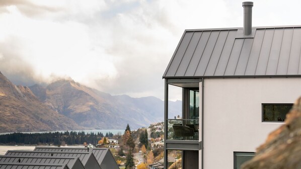 A modern building with a large balcony, a metal roof, and a view of mountains and a lake.