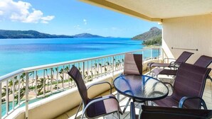 Un balcón con una mesa y sillas de vidrio, con vista a una playa y aguas azul turquesa.