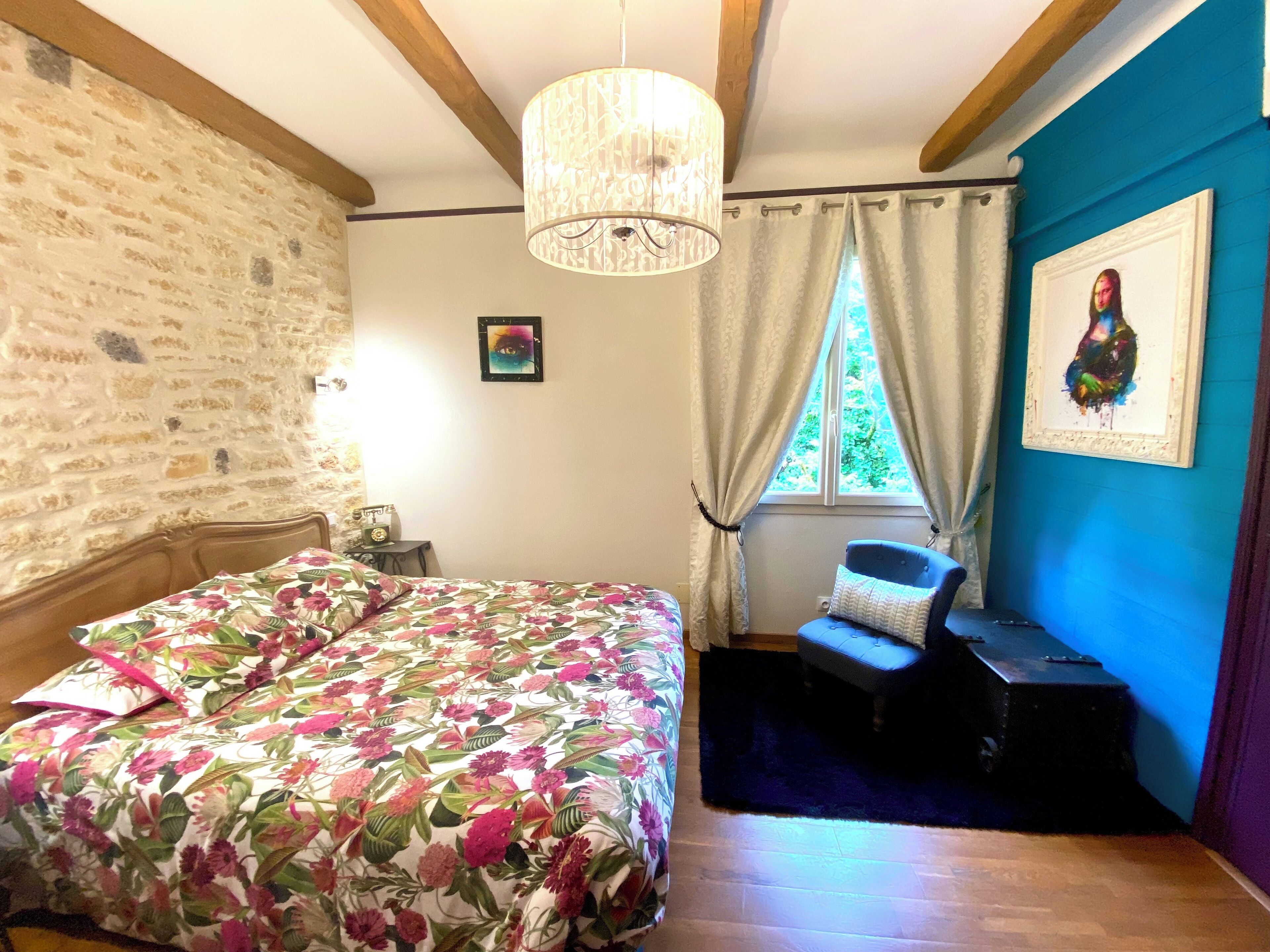 A bedroom with a floral bedspread, a wooden headboard, a chandelier, a window with curtains, a blue wall, and a framed picture.