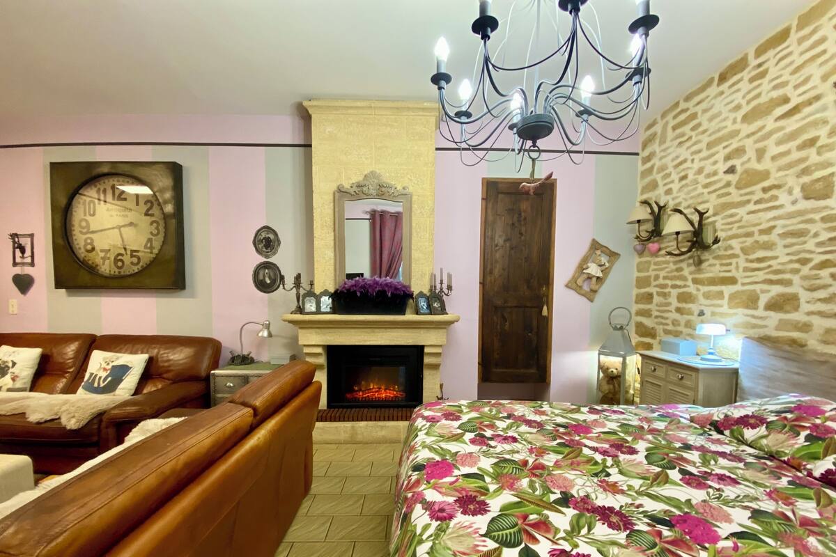 Een knus ingerichte kamer met een open haard, een kroonluchter, een bed met bloemenbedrukking, een leren bank en een klok aan de muur.