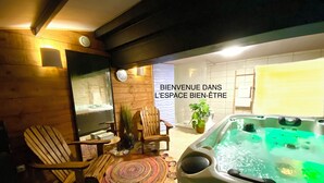 A well-lit indoor space with a hot tub, wooden chairs, a small table, and a decorative rug. - Ô Clair de Lune - Espace bien être avec Spa - Piscine chauffée (Sarlat-la-Canéda)