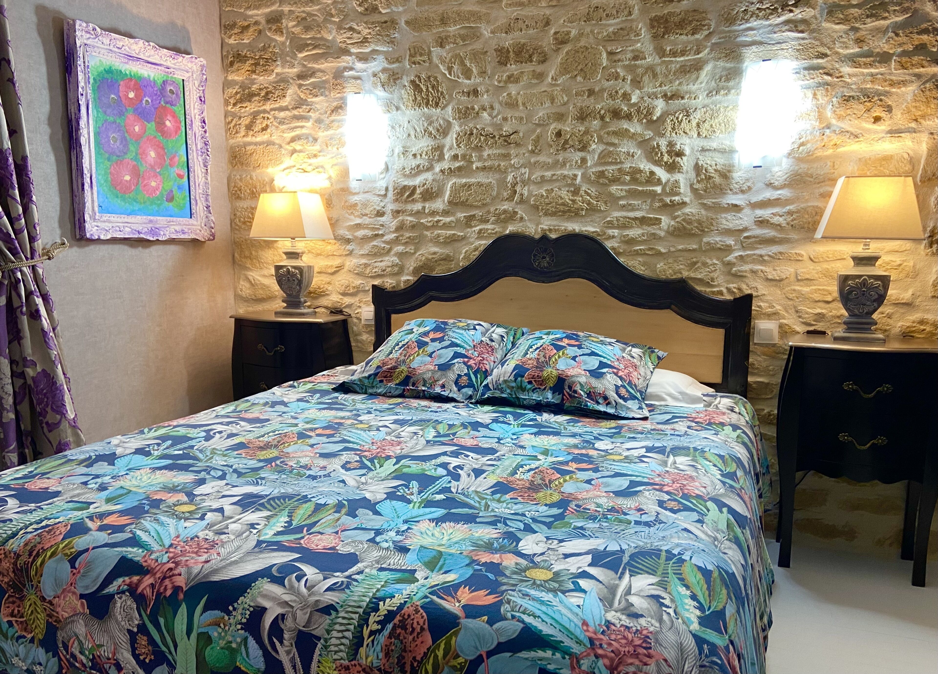 Een slaapkamer met een stenen muur, een bloemenbedekker, twee nachtlampjes en een lijst met een afbeelding aan de muur.