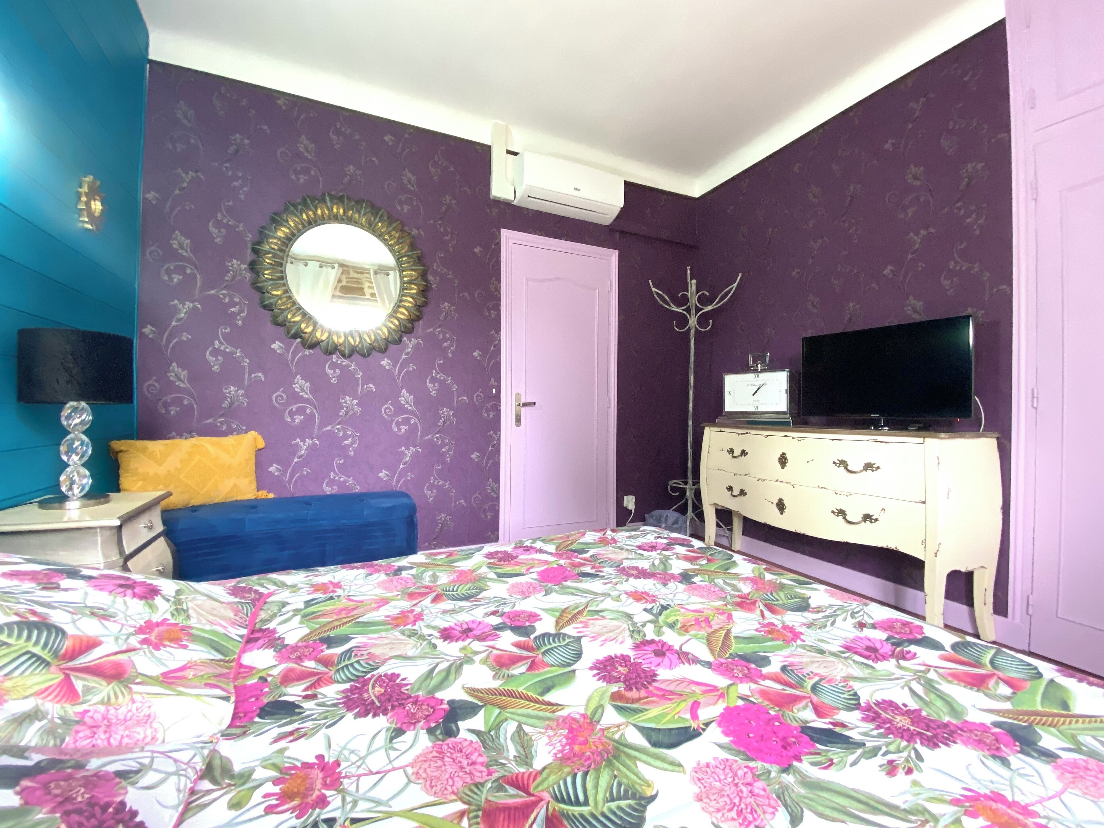 Un dormitorio con colcha estampada, televisor en una cómoda, espejo en la pared y una puerta.