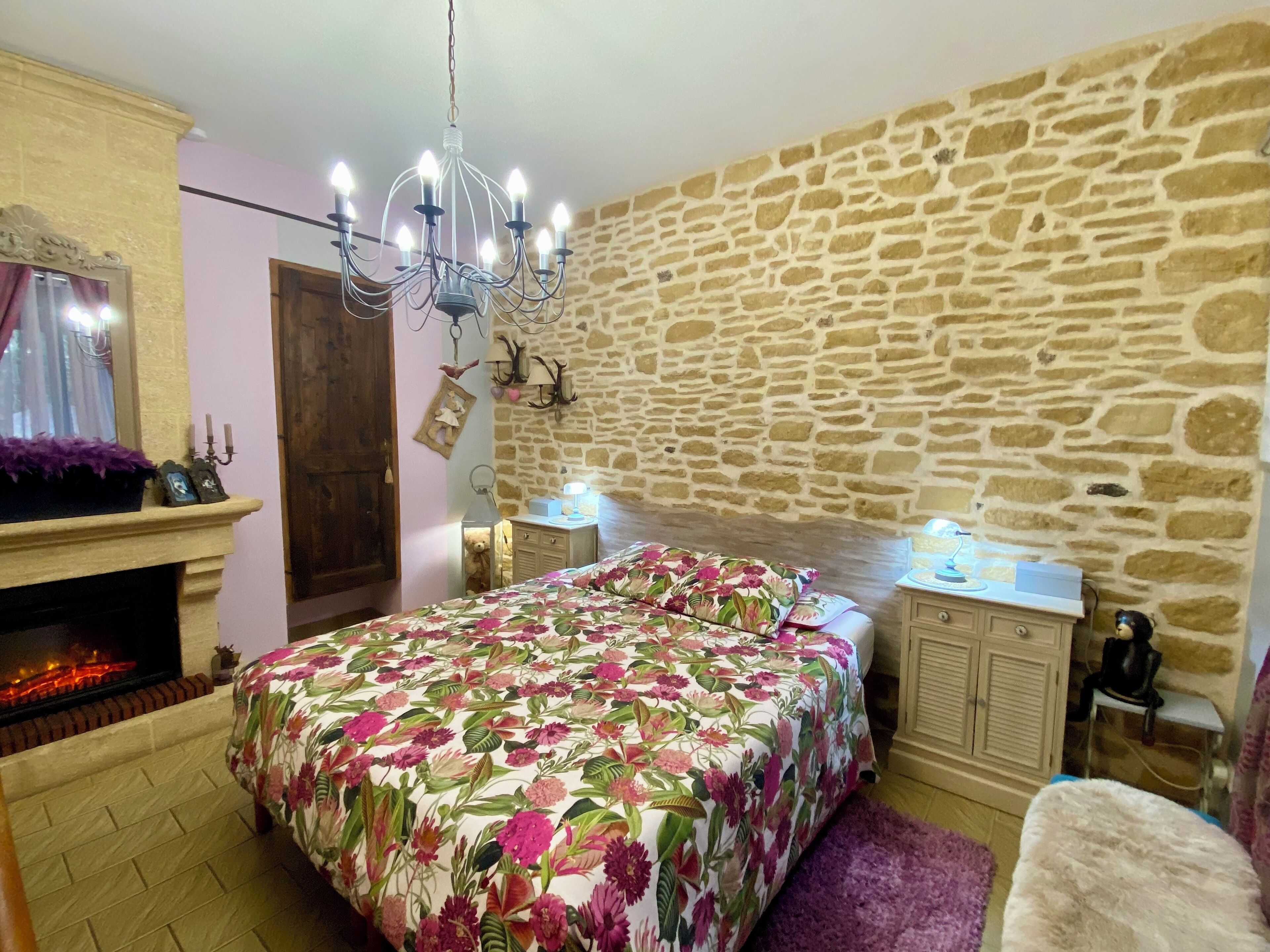 A bedroom with a stone wall, a chandelier, a bed with floral bedding, a fireplace, and bedside lamps.