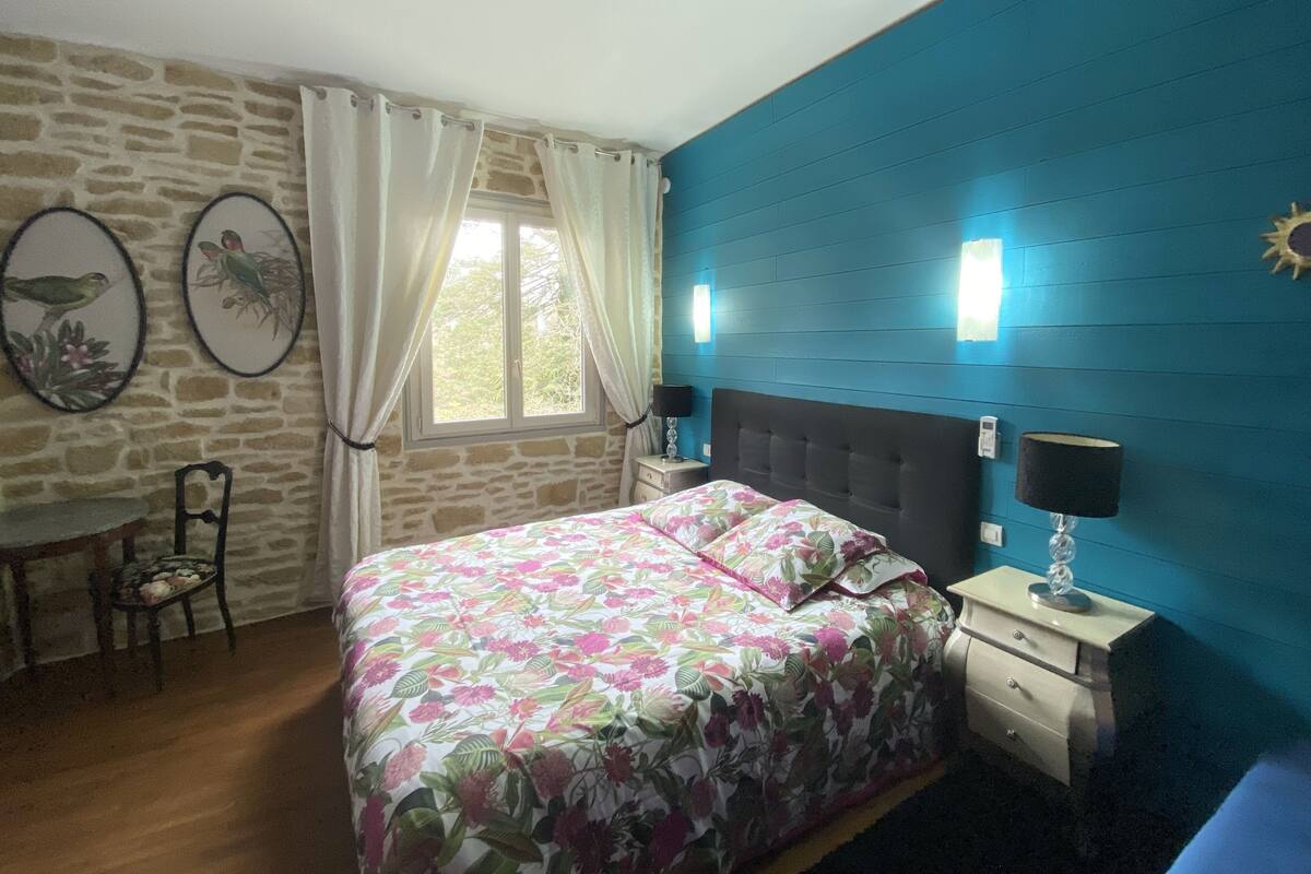 Een slaapkamer met een bed, nachtkastje, stoel, raam met gordijnen en wandlampen.