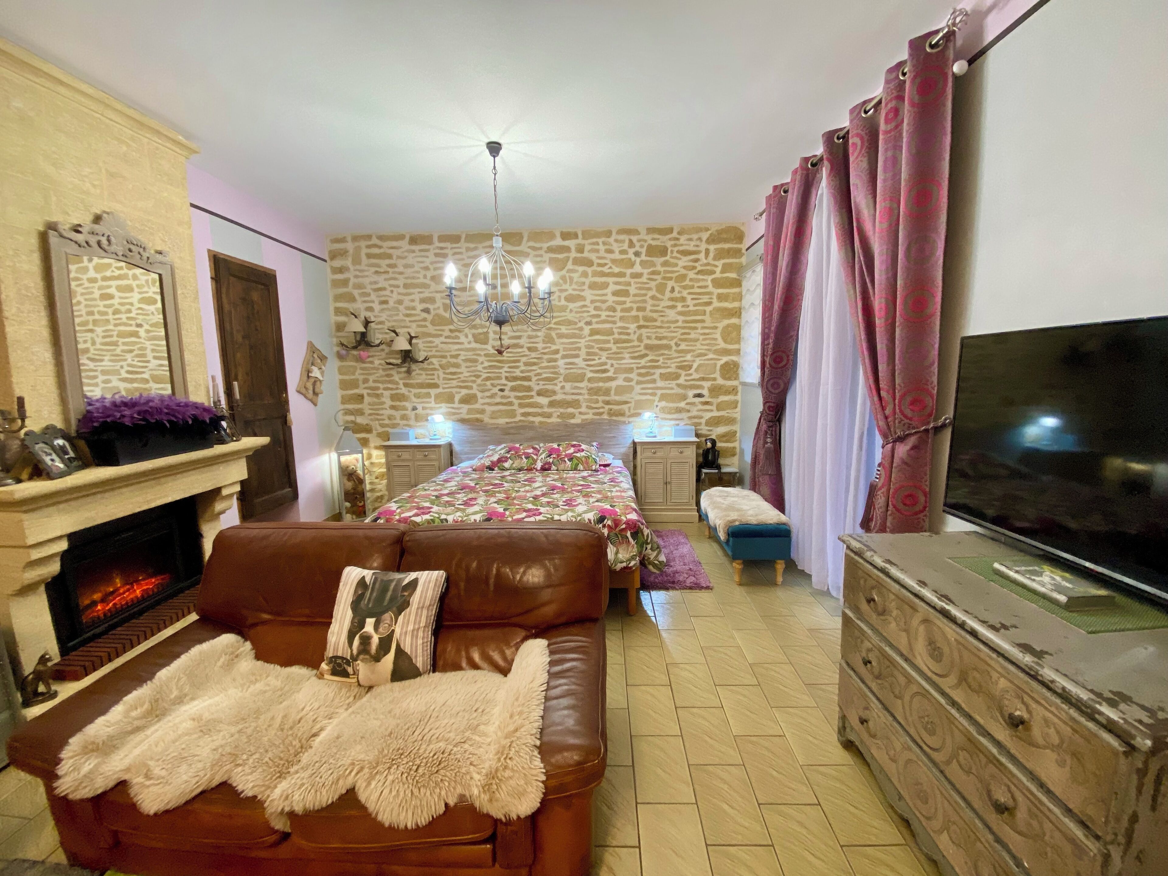A cozy bedroom with a stone wall, a fireplace, a bed with floral bedding, a leather sofa, a TV, and a dresser.