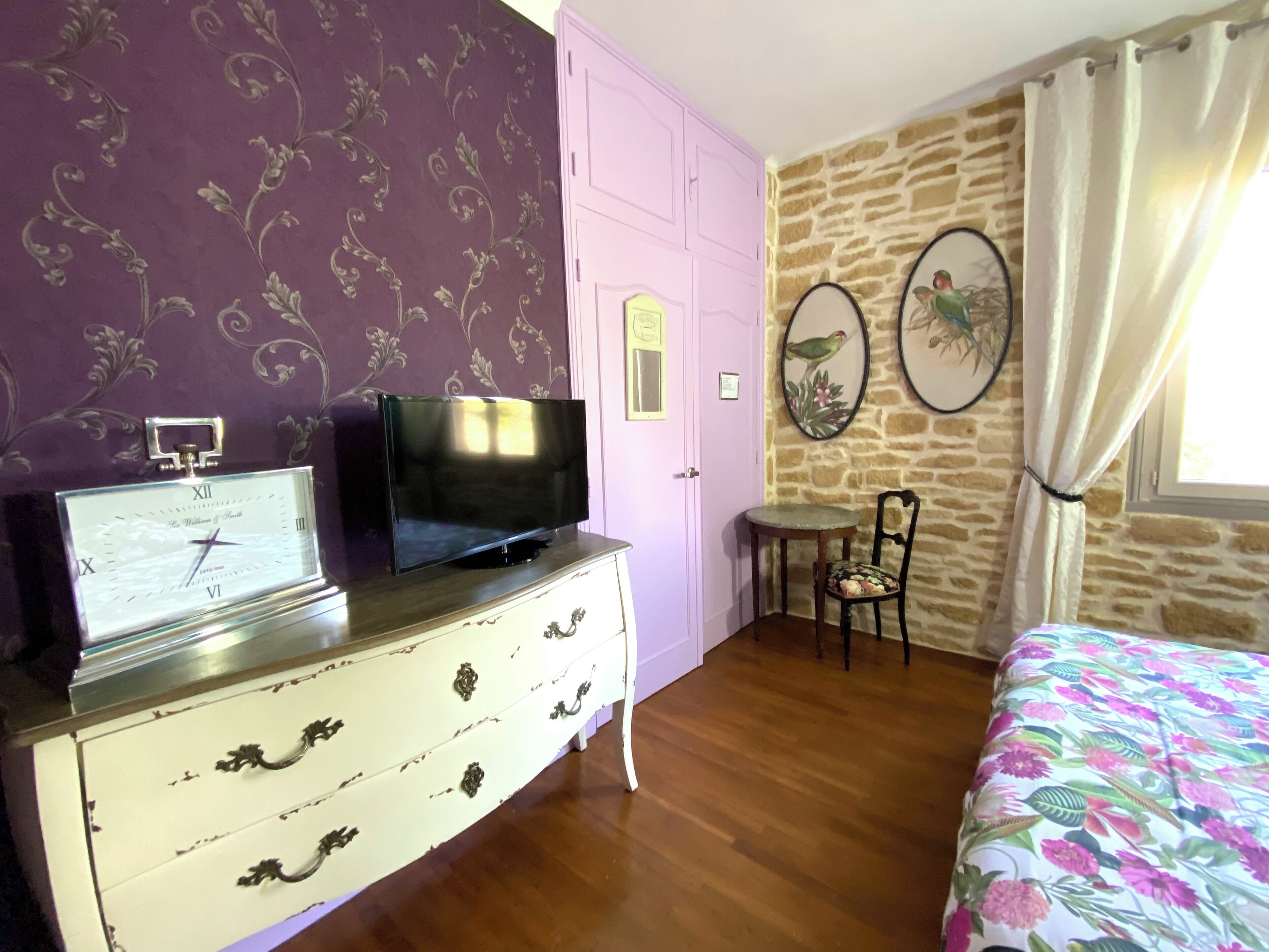 Ein Zimmer mit Holzboden, einem weißen Schminktisch mit Uhr, einem Fernseher, einer rosafarbenen Tür, einem kleinen Tisch mit Stuhl und einem Bett mit blumigem Überwurf.