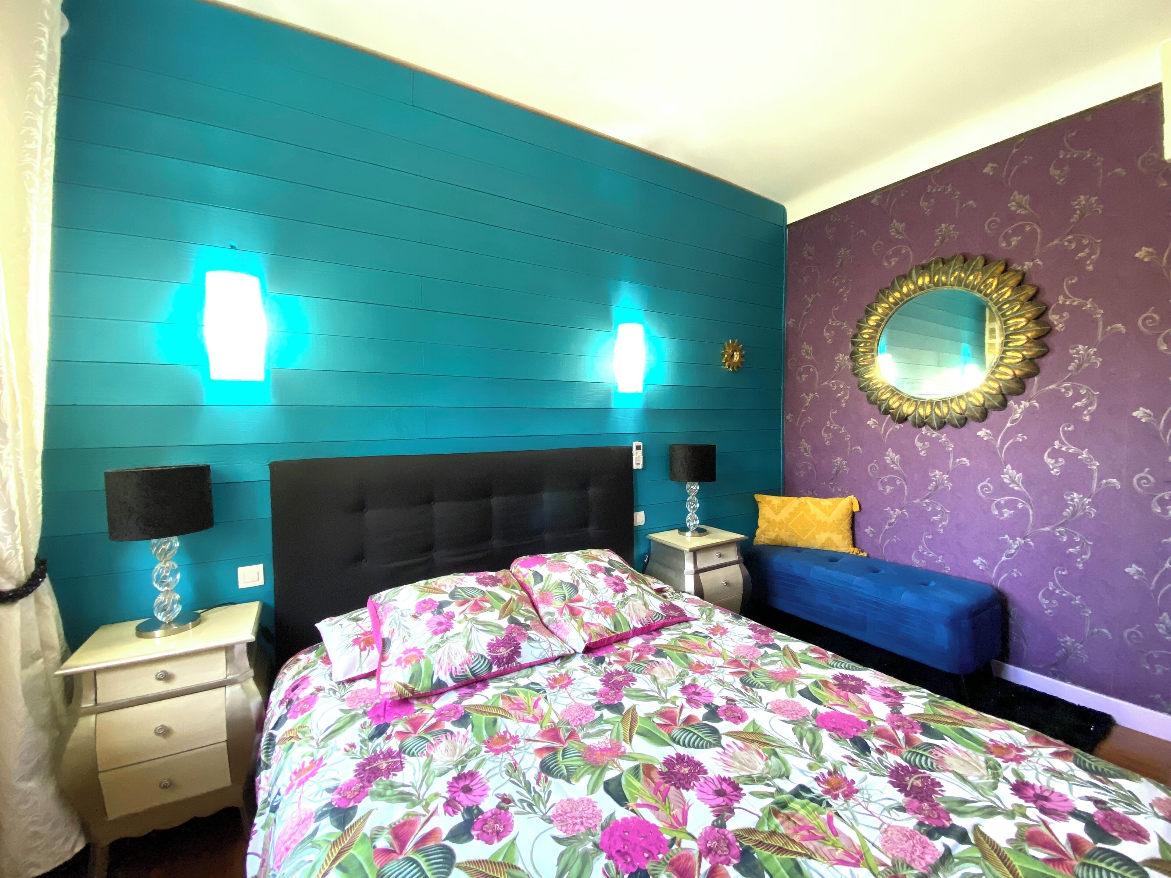 A bedroom with teal walls, a floral bedspread, a blue sofa, and a decorative mirror.