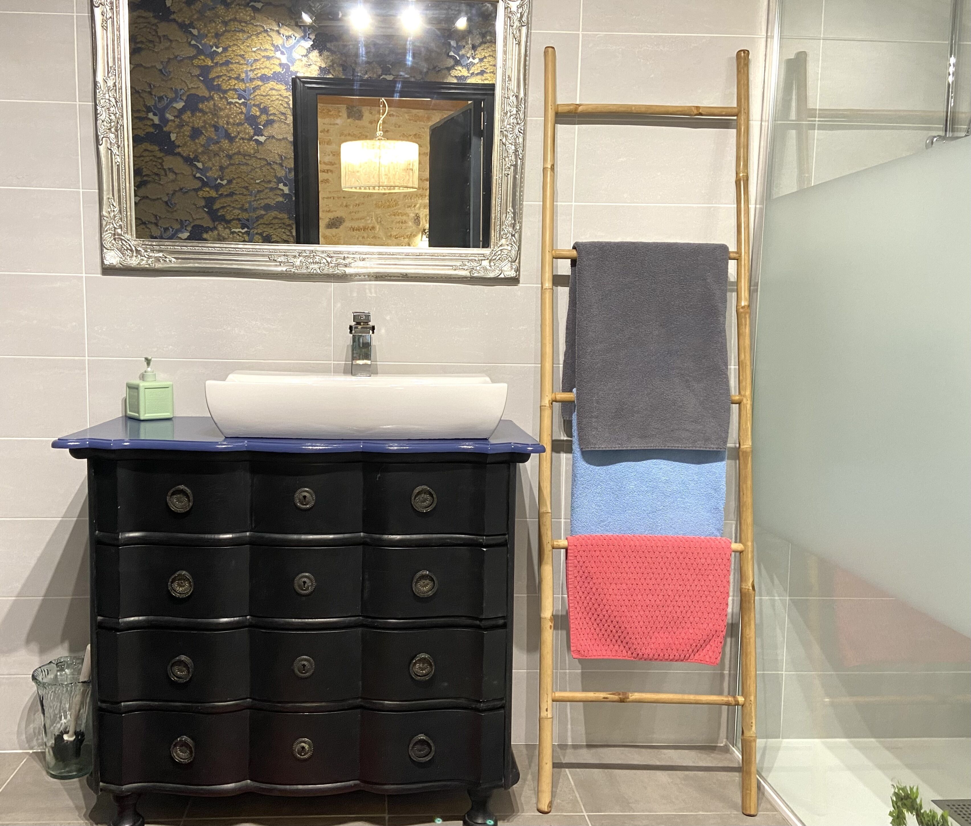 Un baño con tocador negro, un espejo, un lavamanos y un perchero con tres toallas.