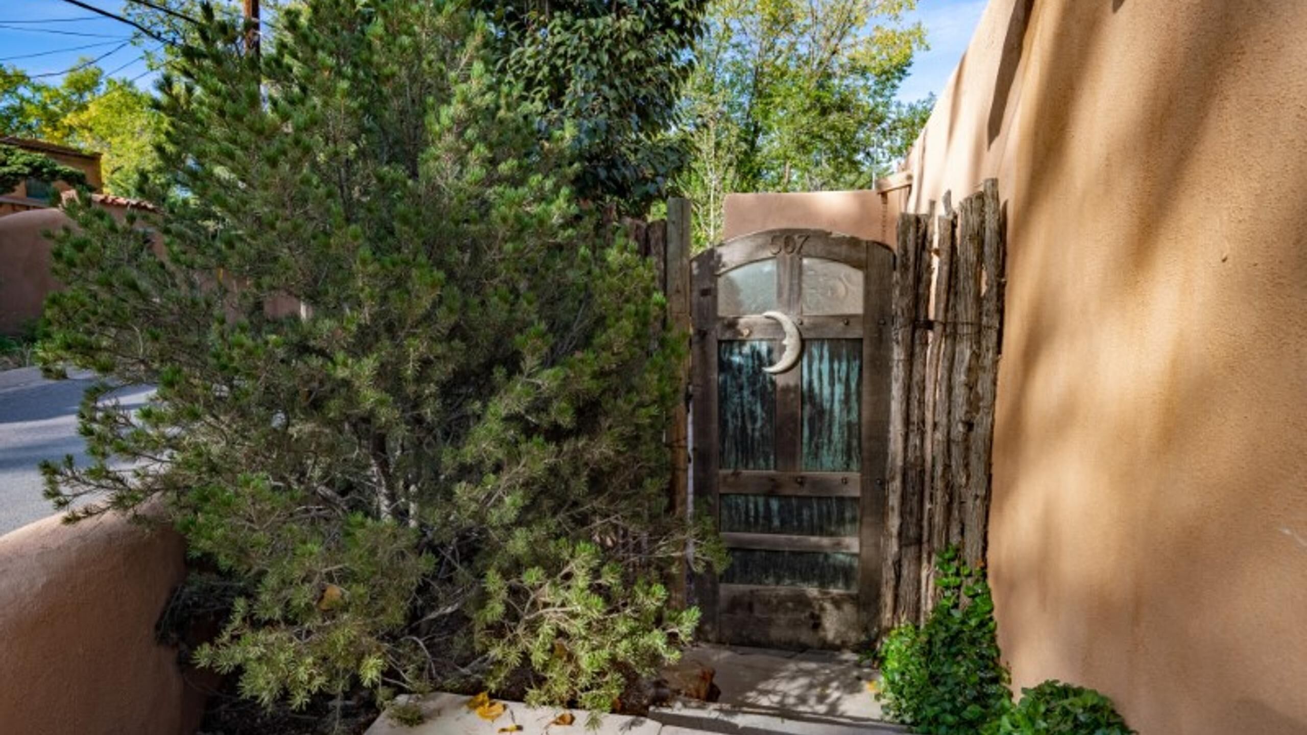 A rustic wooden gate with a metal plaque, surrounded by dense greenery and a building with a stucco exterior.