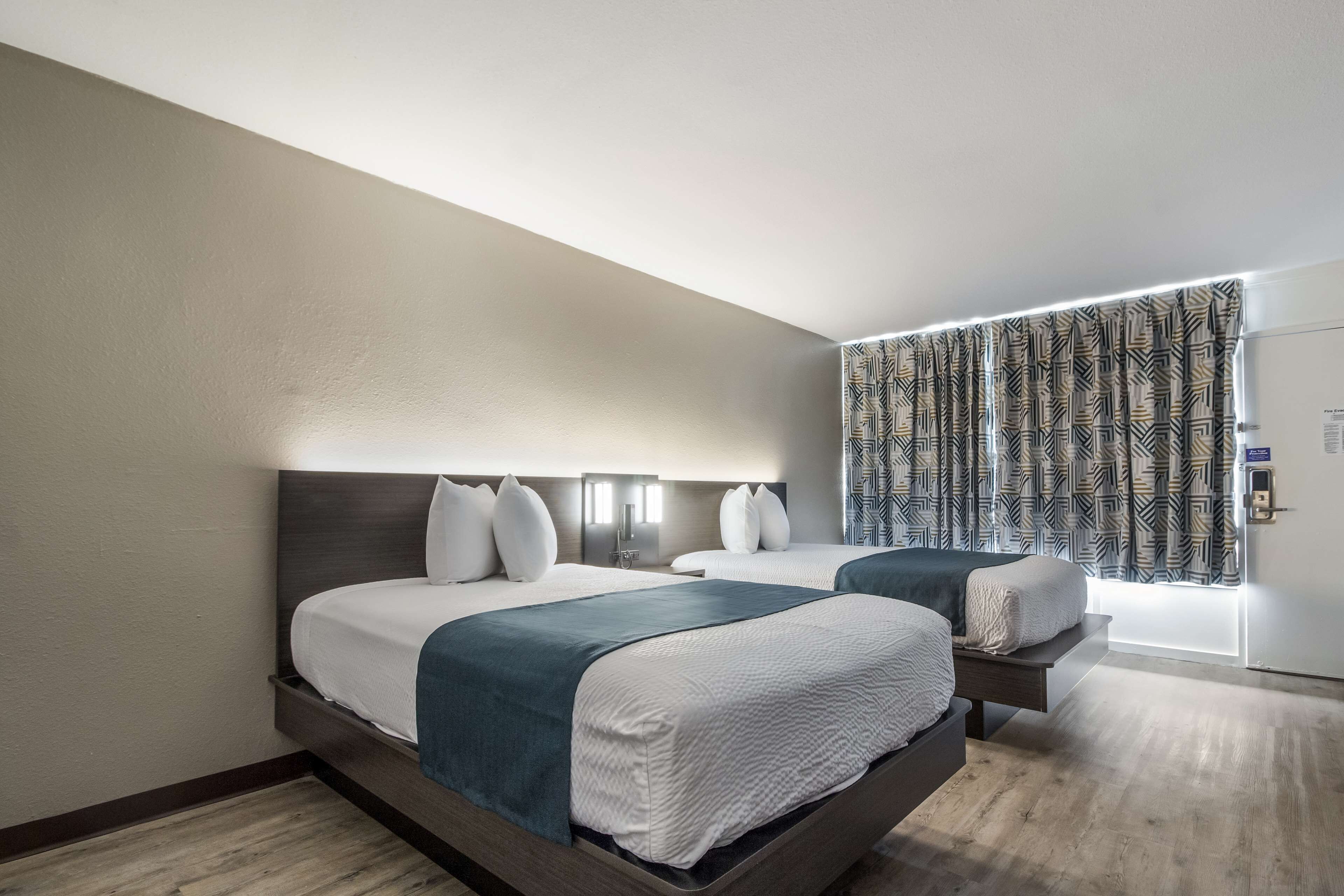 A modern hotel room with two beds, a large window with patterned curtains, and a wooden floor.