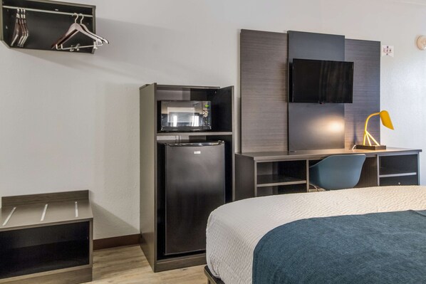 A hotel room with a bed, a desk, a chair, a TV, a refrigerator, and a coat rack.