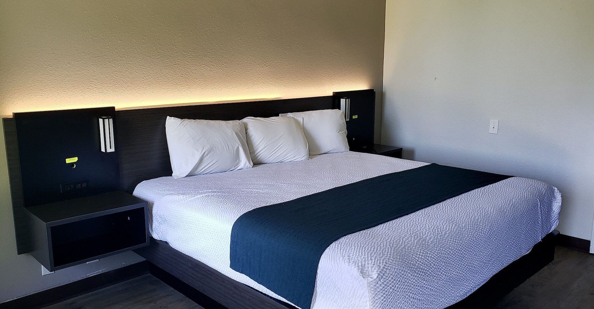 A neatly made bed with white linens and a dark blue throw blanket, flanked by two bedside tables with black surfaces and a headboard with integrated lighting.