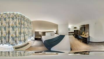 Una habitación de hotel moderna con techo curvo, una cama grande con una manta oscura, un escritorio con silla y un ventanal con cortinas estampadas.