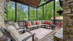 An outdoor covered patio with a stone wall, a wooden ceiling, and a large window overlooking a forest.