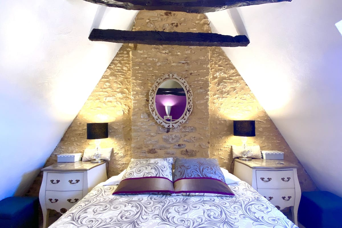 A bedroom with a stone wall, a bed with patterned bedding, two bedside tables with lamps, and a mirror on the wall.