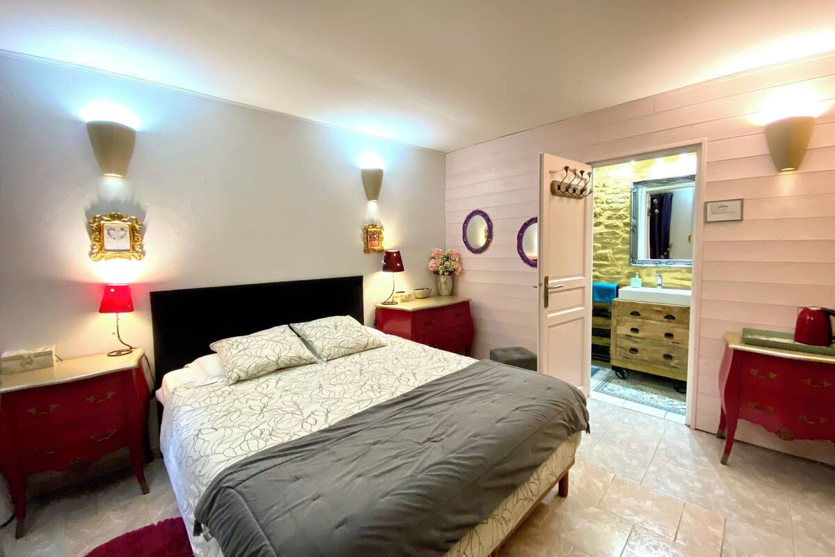 A bedroom with a bed, two bedside tables, a nightstand, a red dresser, a mirror, and a door leading to a bathroom.