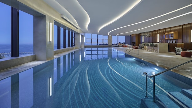 A modern indoor swimming pool with a view of the ocean, a bar area, and seating. - Hard Rock Hotel Dalian (Dalian)