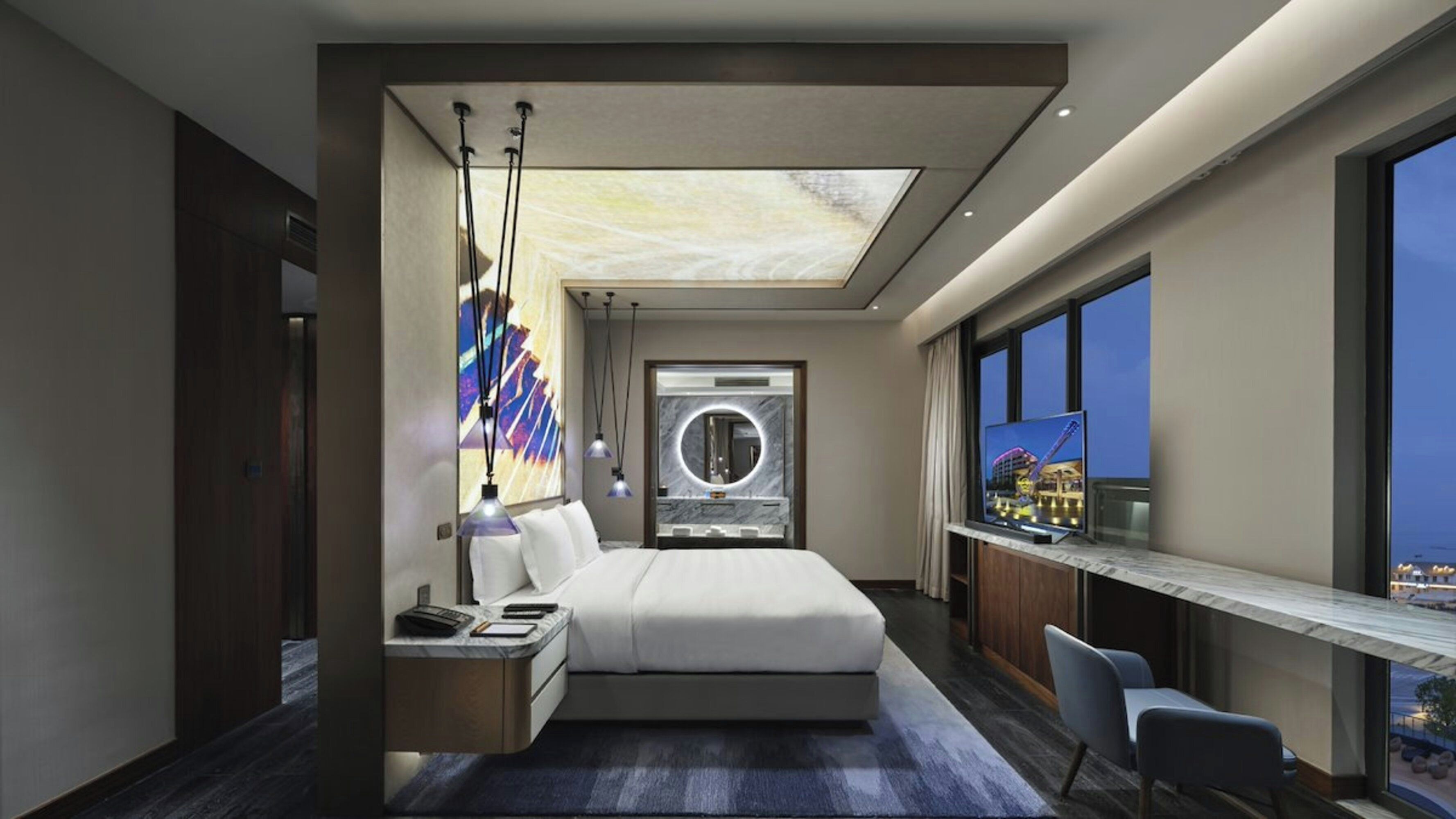 Una habitación de hotel moderna con una cama grande, un escritorio con computadora, un ventanal con vistas y un espejo circular sobre la cama.