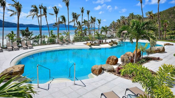 A swimming pool with lounge chairs, palm trees, and a view of the ocean. - Whitsunday Apartment West 1002 (Hamilton Island)
