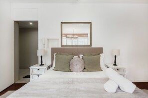 A neatly arranged bedroom with a large bed, two bedside tables, a mirror, and a door leading to another room.