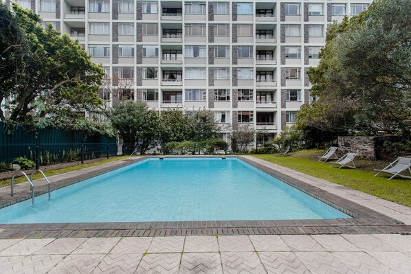 A rectangular swimming pool with a tiled border, surrounded by a paved area with lounge chairs and a building with multiple balconies in the background.