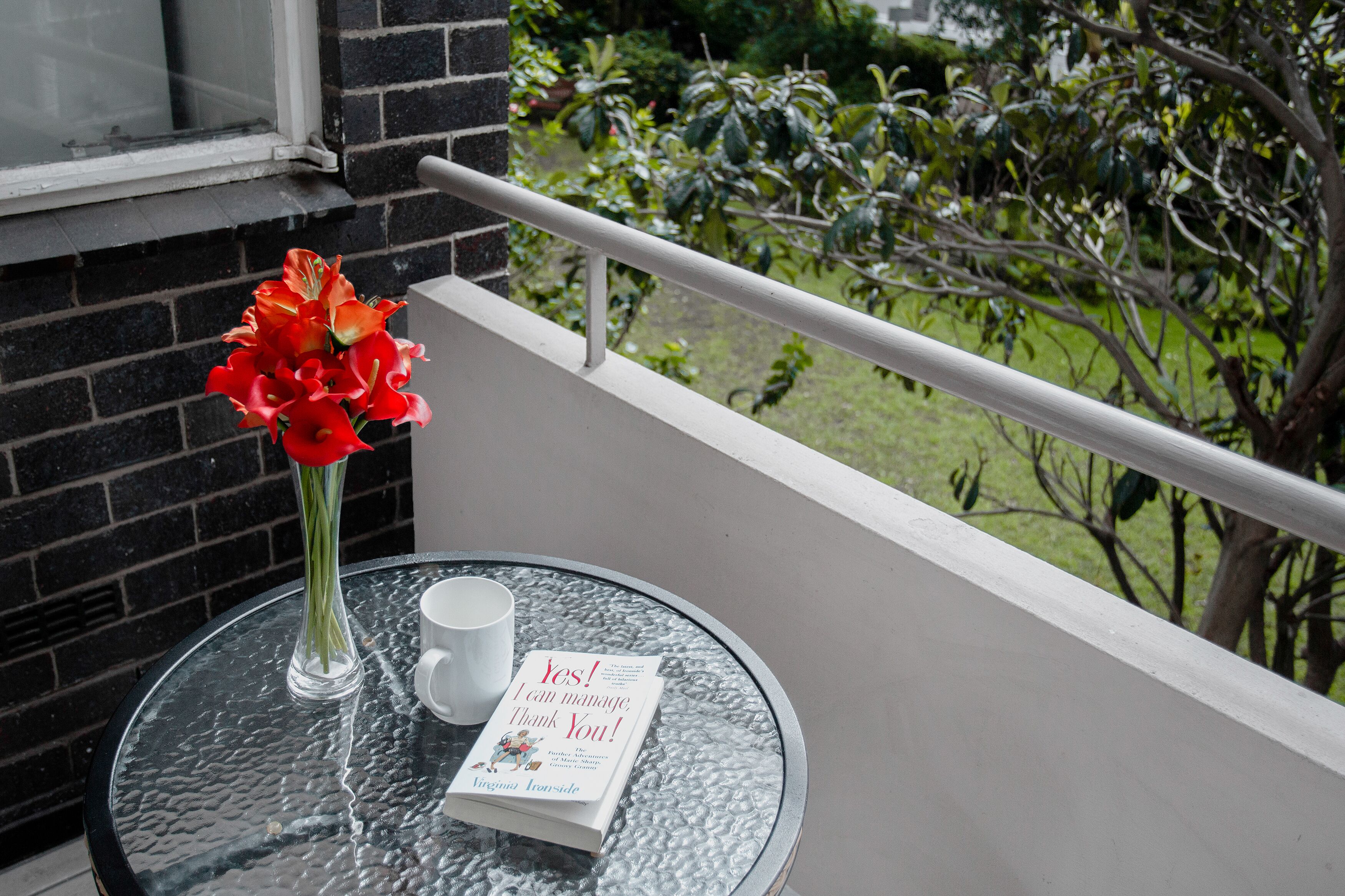 A round glass table with a white mug and a book titled "Yes! I can manage, Thank You!" on a balcony with a view of greenery.