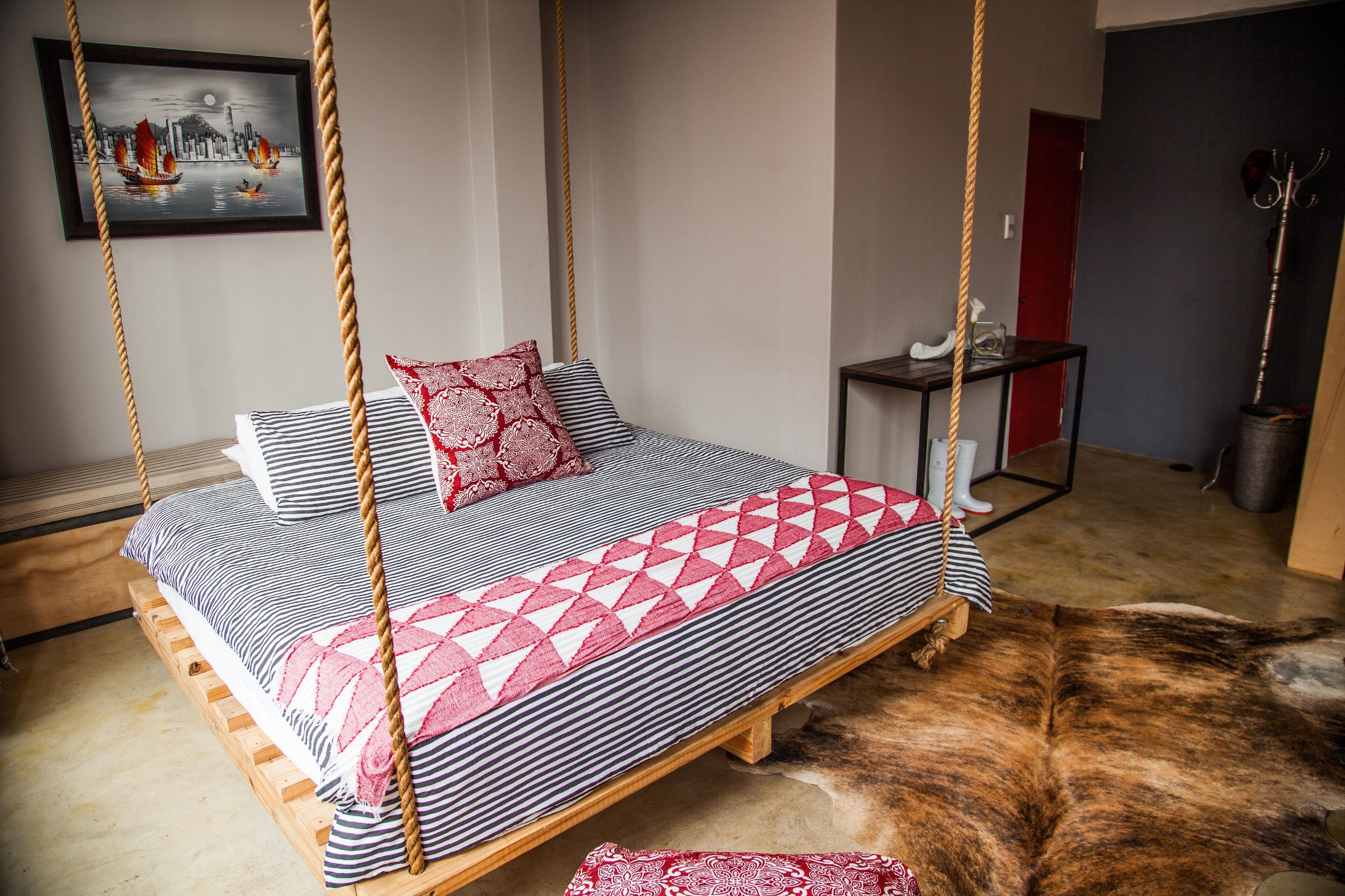 A bed with a geometric patterned bedspread, a red and white pillow, and a striped pillow. There is a small table with a lamp and a coat rack. A framed picture hangs on the wall.