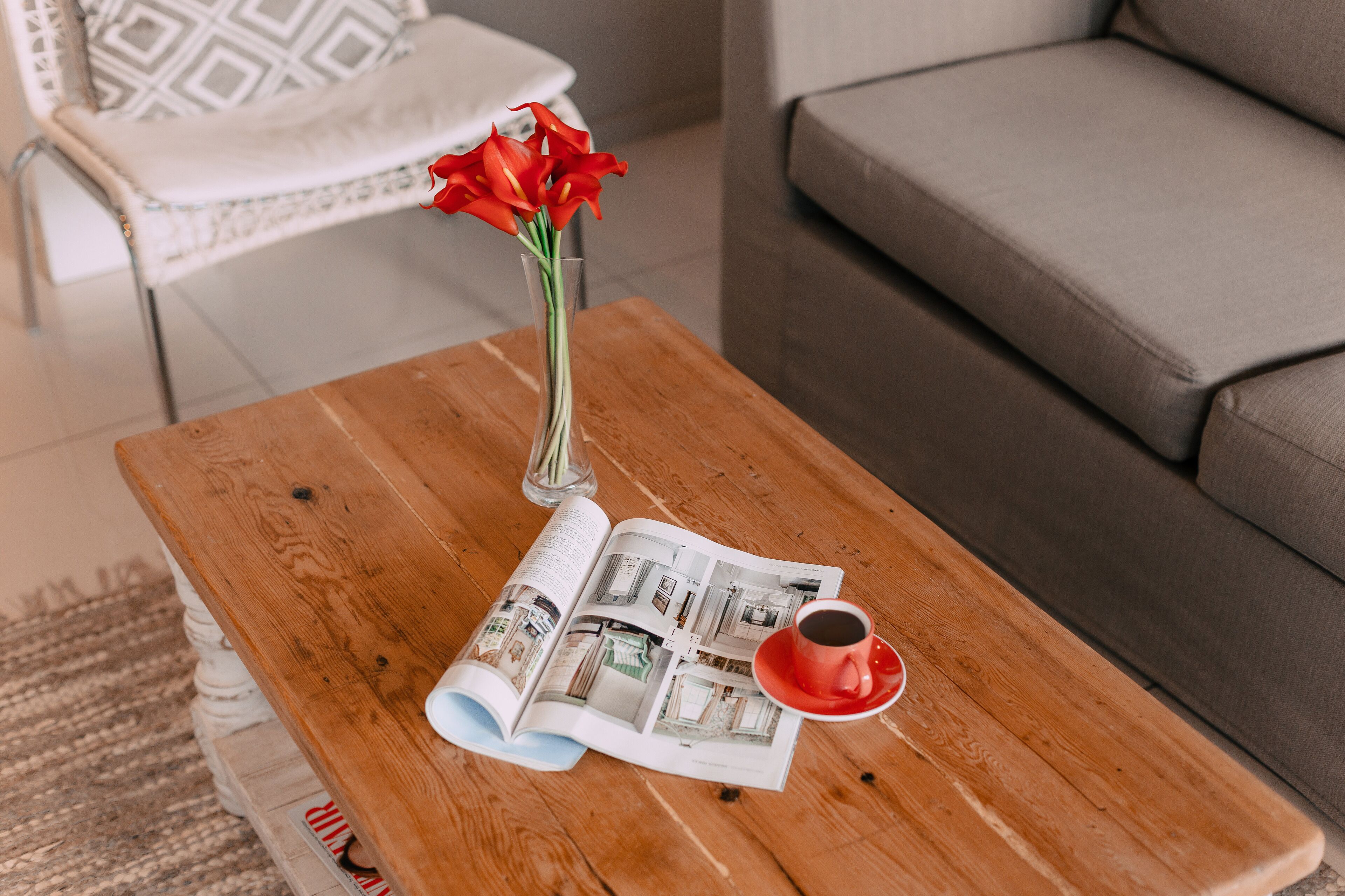 Una mesa de madera con una revista, una taza roja y un florero con flores rojas.