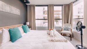 Une chambre d’hôtel avec un grand lit, une chaise, un ventilateur sur pied et une vue sur la ville par les fenêtres.