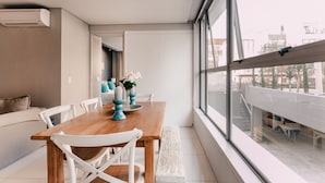 Un espace de restauration moderne avec une table en bois, des chaises blanches et une vue sur la ville grâce à de grandes fenêtres.