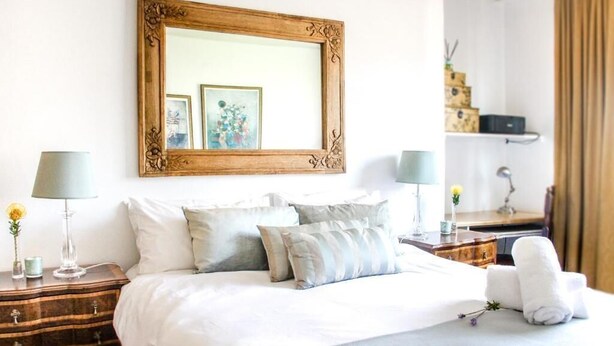 A neatly made bed with white linens and multiple pillows, a wooden nightstand with a lamp, and a framed mirror on the wall. - 438 St Martini (Cape Town)
