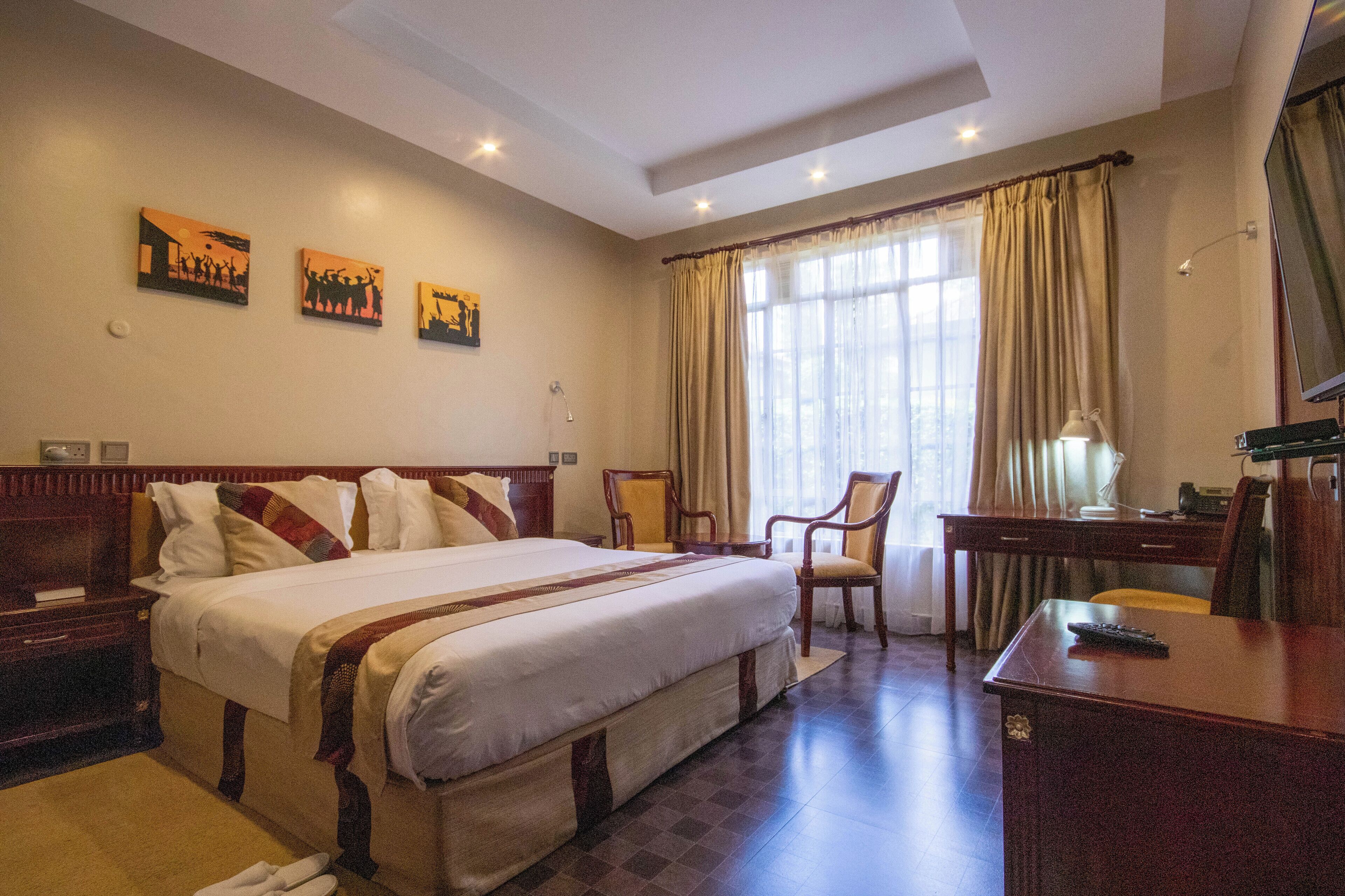 A hotel room with a large bed, a desk, a chair, and artwork on the wall.