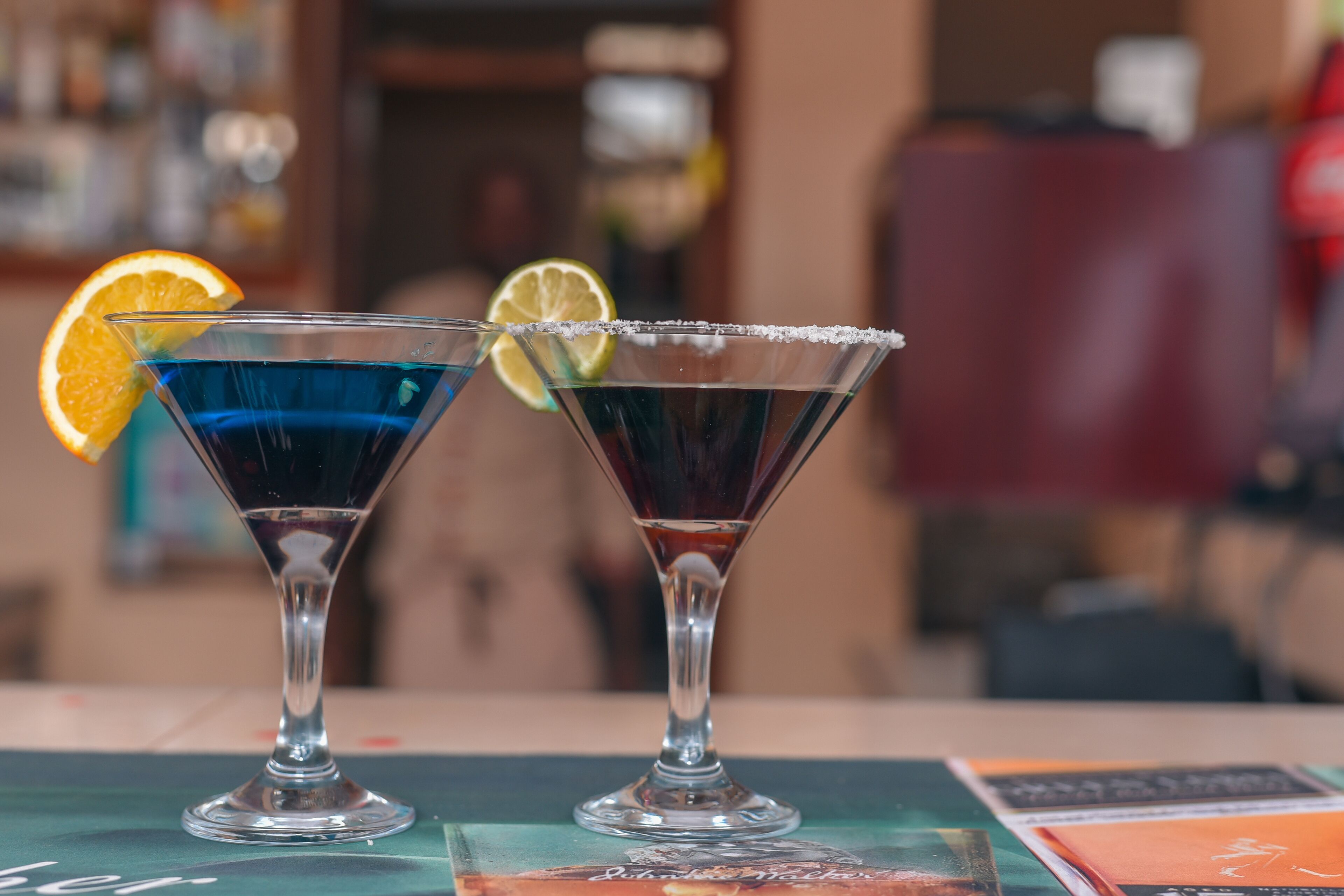 Two martini glasses with a blue drink and a dark drink, garnished with an orange slice and a lime wedge, on a table with a menu.